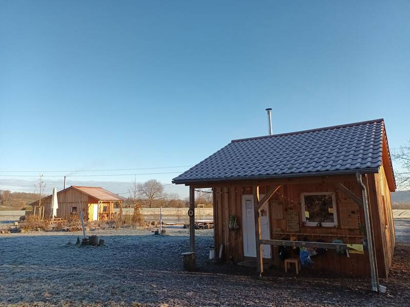 Zwei kleine Holzhütten stehen in einer frostigen, ländlichen Landschaft unter klarem, blauem Himmel.