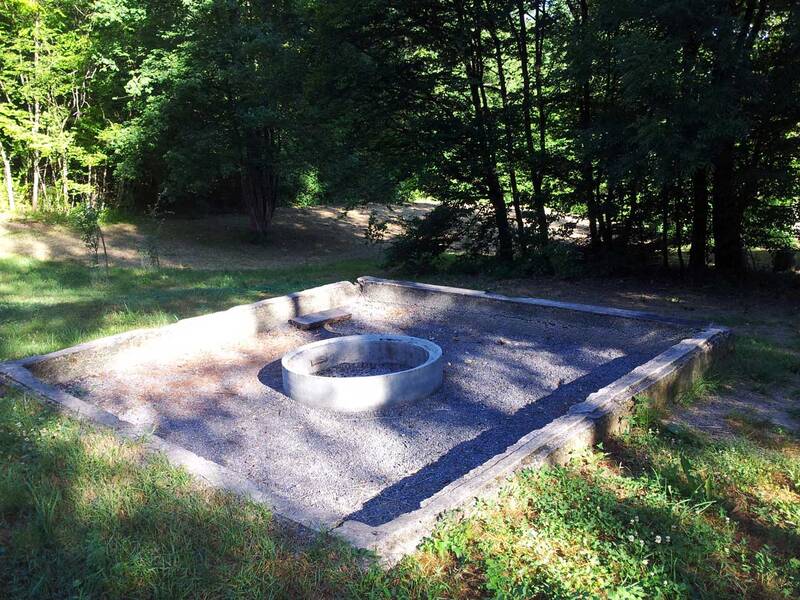 Eine kreisförmige Feuerstelle aus Beton befindet sich in der Mitte eines rechteckigen, mit Kies bedeckten Platzes in einem Waldgebiet.