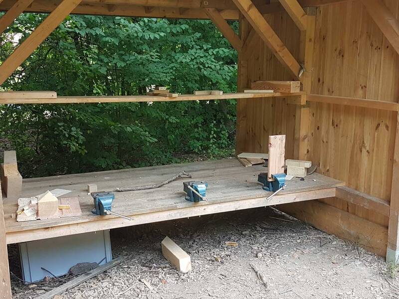 Eine Werkbank unter einer Holzüberdachung im Wald, drei Schraubstöcke sind zu sehen.