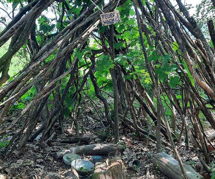 Ein Tipi aus Holzstöcken steht im Wald, mit einem Schild, auf dem "Feuerwache" steht, und verschiedenen Gegenständen wie Siebe und Holzstümpfe am Boden.