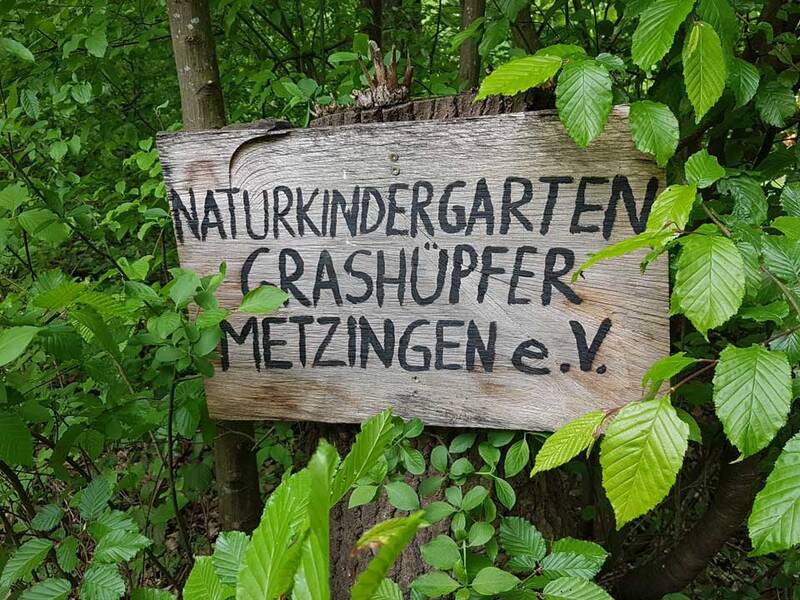 Ein handgemachtes Holzschild mit der Aufschrift "Naturkindergarten Crashüpfer Metzingen e.V." hängt zwischen grünen Blättern an einem Baum.