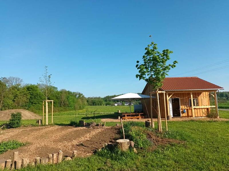 Eine kleine Holzhütte mit rotem Dach steht inmitten einer grünen Landschaft, flankiert von jungen Bäumen und einem weißen Sonnenschirm.