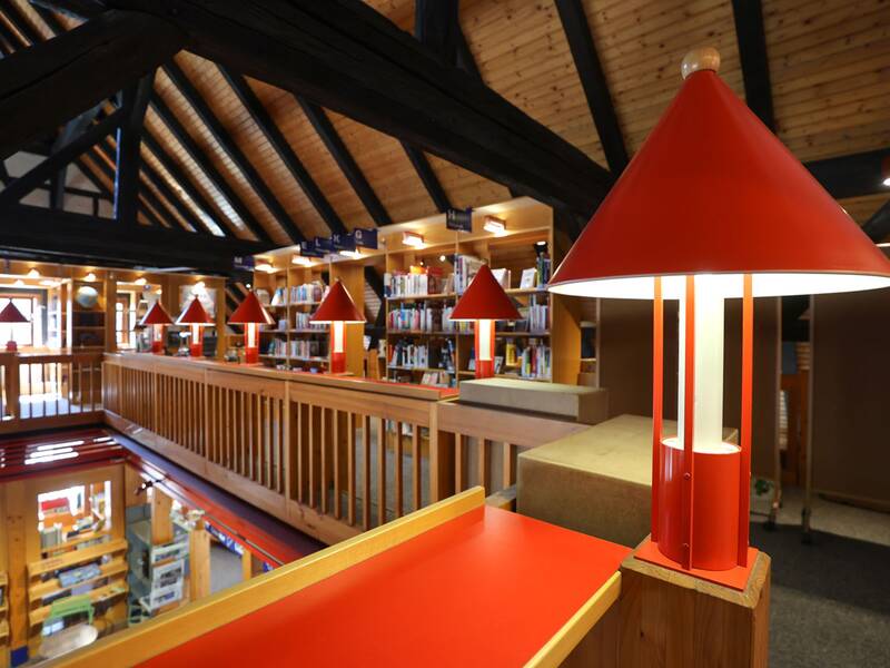 Stadtbücherei, Eine Bibliothek mit einem offenen Zwischengeschoss, eingerahmt von Holzbalken und gesäumt von mehreren roten Tischlampen.