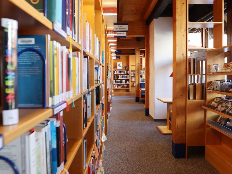 Stadtbücherei, Ein langer Gang in einer Bibliothek ist von hölzernen Bücherregalen gesäumt, die mit zahlreichen Büchern gefüllt sind.
