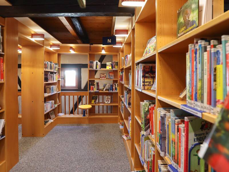 Stadtbücherei, Eine gemütliche Bibliothek mit Holzbücherregalen, die dicht mit Büchern gefüllt sind, darunter ein kleiner Holztisch und ein Fenster im Hintergrund.