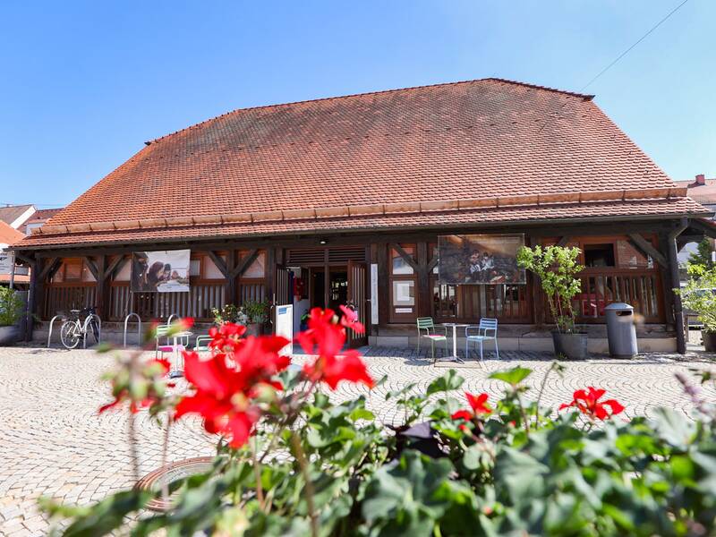 Stadtbücherei, Ein traditionelles, hölzernes Gebäude mit rotem Ziegeldach und mehreren Stühlen davor, umgeben von Blumentöpfen und gepflastertem Boden, bei sonnigem Wetter.