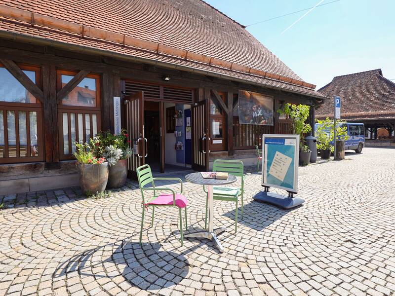 Stadtbücherei, Vor dem Fachwerkgebäude mit einem offenen Eingang stehen zwei bunte Stühle und ein Tisch auf einem gepflasterten Platz.