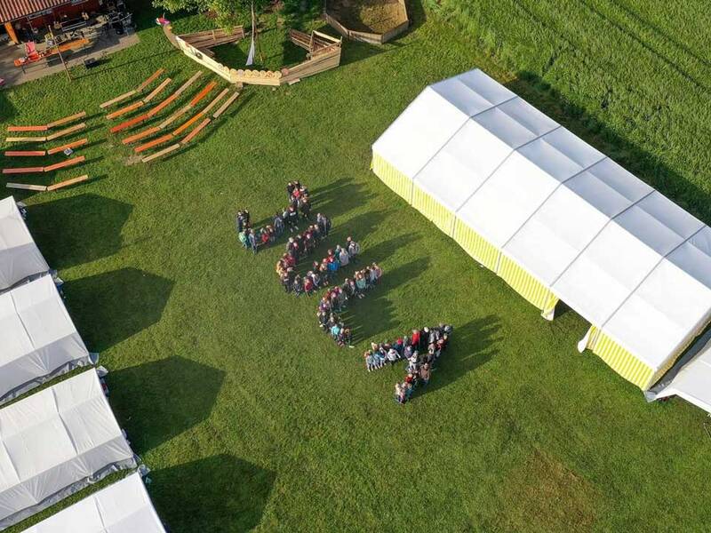 Eine Gruppe von Menschen steht in Form eines Fragezeichens auf einer Wiese, umgeben von weißen Zelten und Bänken.