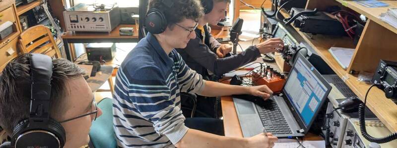 Drei Personen mit Kopfhörern sitzen in einem Technikraum vor Computern und Funkgeräten und arbeiten konzentriert.
