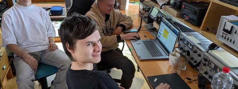 Drei Personen sitzen in einem Raum mit technischen Geräten und Computern, während eine Person auf einem Laptop arbeitet.