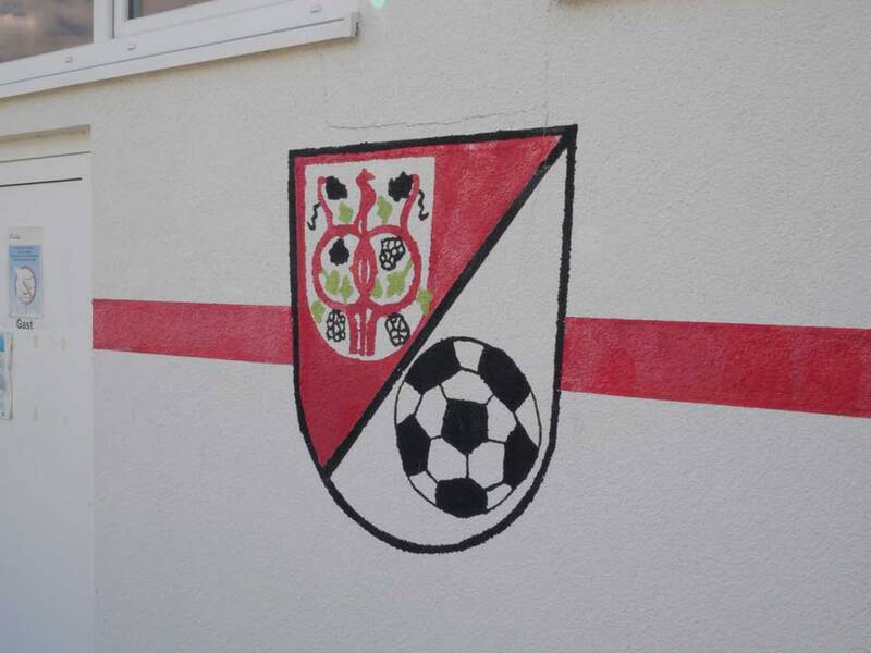 Das Bild zeigt eine Wand mit einem Wappen, das in Rot, Schwarz und Weiß gemalt ist und ein Schild mit Verzierungen sowie einen Fußball enthält.