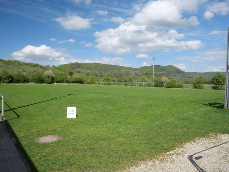 Ein grüner Sportplatz mit einem Schild, auf dem "Platz gesperrt" steht, ist von Hügeln und einem blauen Himmel mit Wolken umgeben.