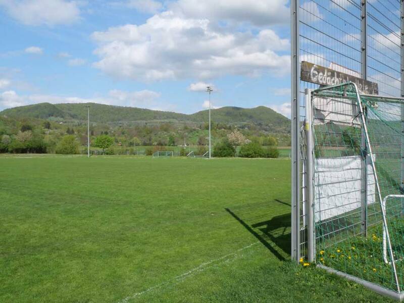 Ein grünes Fußballfeld mit einem Tor im Vordergrund, umrahmt von einer hügeligen Landschaft unter einem blauen Himmel mit einigen Wolken.