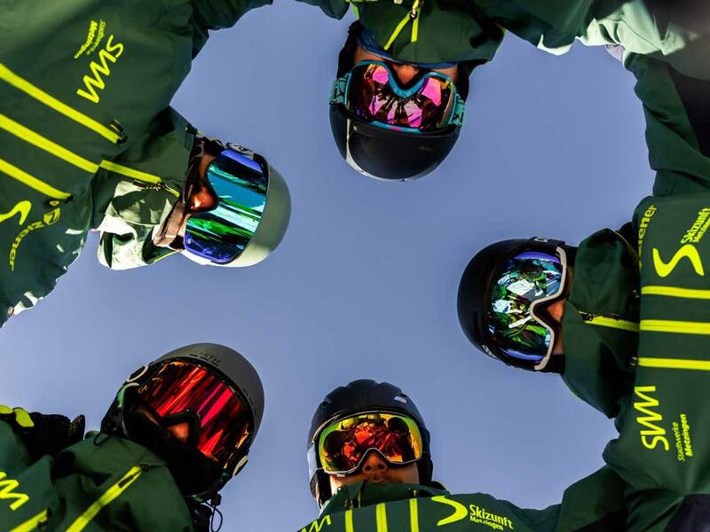 Fünf Personen in grünen Winterjacken und Helmen stehen im Kreis und schauen in die Kamera, während der Himmel im Hintergrund zu sehen ist.