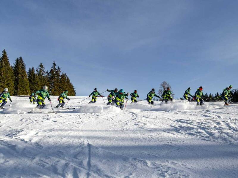 Eine Gruppe von Skifahrern in grüner Kleidung fährt synchron eine Skipiste hinunter, umgeben von schneebedeckten Bäumen.