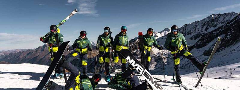 Eine Gruppe von acht Menschen in einheitlicher Skibekleidung posiert mit Skiern und Snowboards auf einem schneebedeckten Berggipfel vor einer beeindruckenden Bergkulisse.