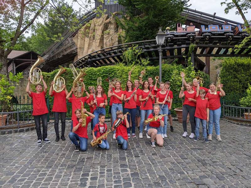 Eine Gruppe von jungen Menschen in roten T-Shirts posiert mit Musikinstrumenten vor einer Achterbahn im Freien.