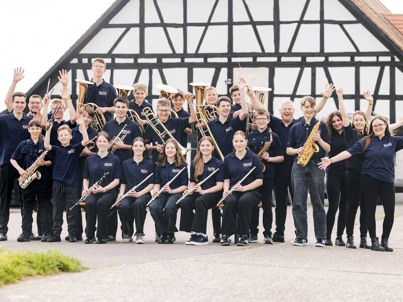 Eine Gruppe junger Musikerinnen und Musiker in einheitlicher Kleidung steht mit verschiedenen Blasinstrumenten vor einem traditionellen Fachwerkhaus und winkt fröhlich in die Kamera.