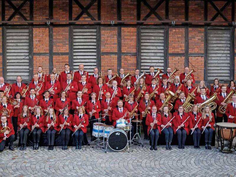 Eine große Blaskapelle in roten Uniformen posiert mit ihren Instrumenten vor einem Ziegelgebäude.