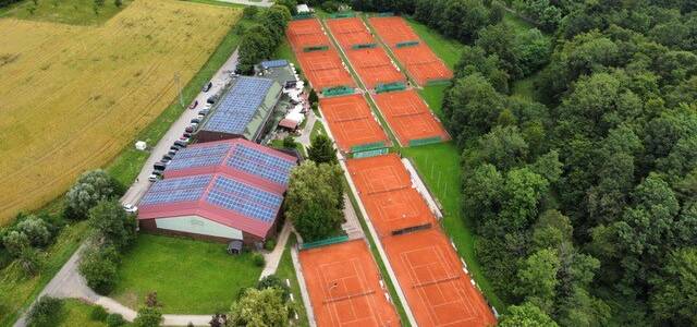 Luftaufnahme einer großen Tennisanlage mit vielen roten Sandplätzen, einem Vereinsgebäude mit Solardach und Wald ringsum.