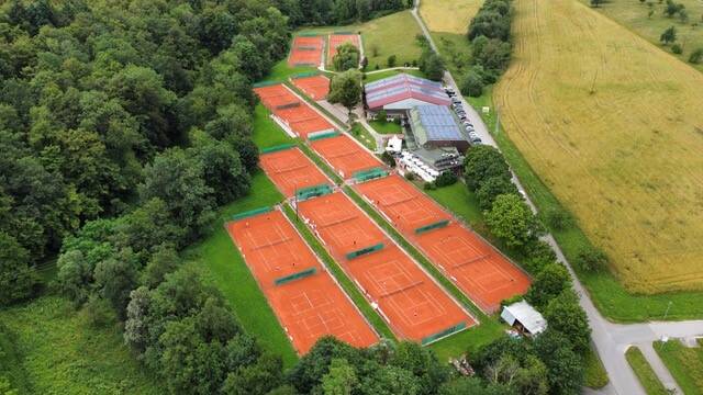 Mehrere parallel angeordnete Tennisplätze liegen eingebettet zwischen Feldern und dichtem Grün, aus der Vogelperspektive gesehen.
