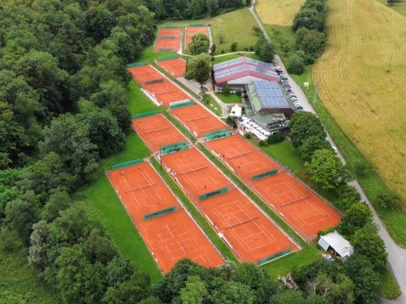Mehrere parallel angeordnete Tennisplätze liegen eingebettet zwischen Feldern und dichtem Grün, aus der Vogelperspektive gesehen.