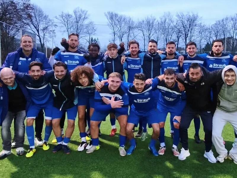 Eine Gruppe von Fußballspielern und Trainern in blauen Trikots posiert lächelnd auf einem Fußballfeld.