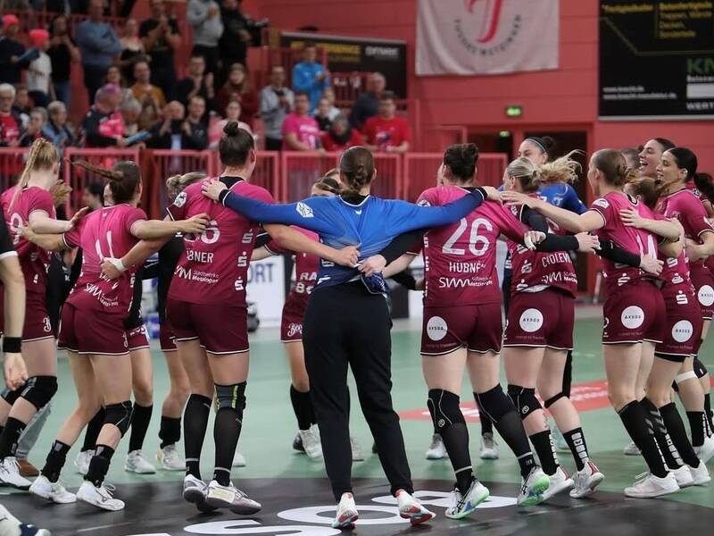 Eine Frauen-Handballmannschaft in weinroten Trikots steht im Kreis auf einem Spielfeld, legt die Arme umeinander und feiert.