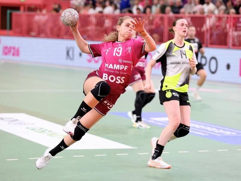 Eine Handballspielerin springt kraftvoll nach vorn und wirft den Ball auf das Tor, während eine Gegenspielerin versucht, sie zu stoppen