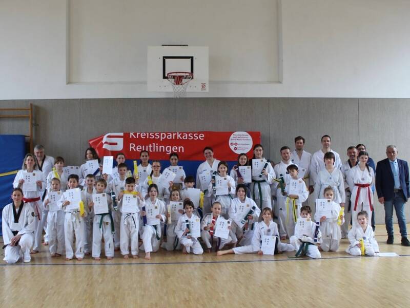 Eine Gruppe von Personen in Taekwondo-Kleidung hält Urkunden vor einer in der Halle aufgestellten „Kreissparkasse Reutlingen“-Banner.