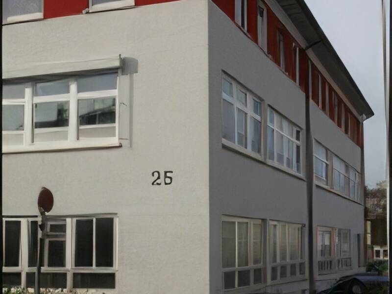 Das Bild zeigt ein modernes, helles Gebäude mit rotem Obergeschoss, vielen Fenstern und der Hausnummer 25 an einer ruhigen Straßenecke.