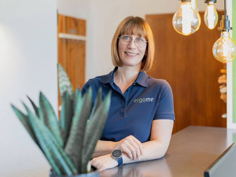 Eine Frau mit Brille und einem dunkelblauen Poloshirt steht lächelnd hinter einem Tresen, umgeben von modernen, hängenden Glühbirnen.