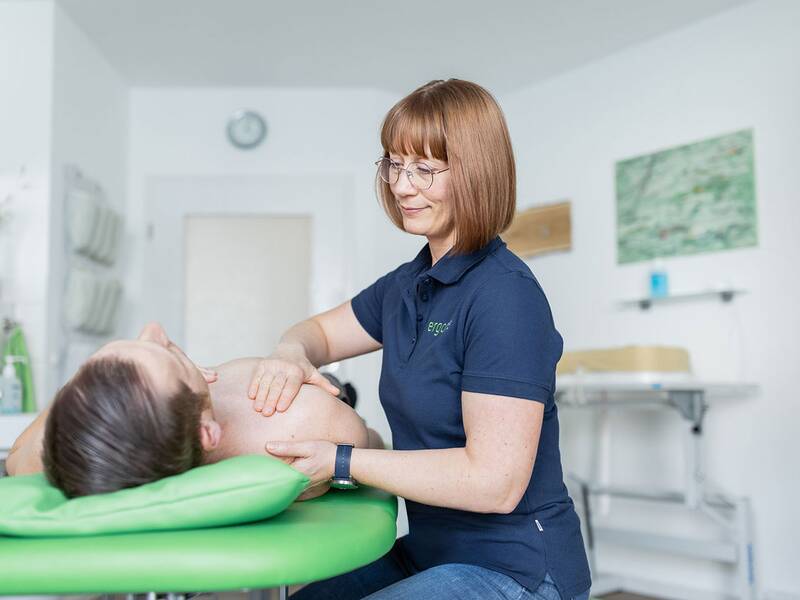 Eine Frau in einem blauen Poloshirt massiert in einem hellen Raum den Rücken einer auf einer Liege liegenden Person.