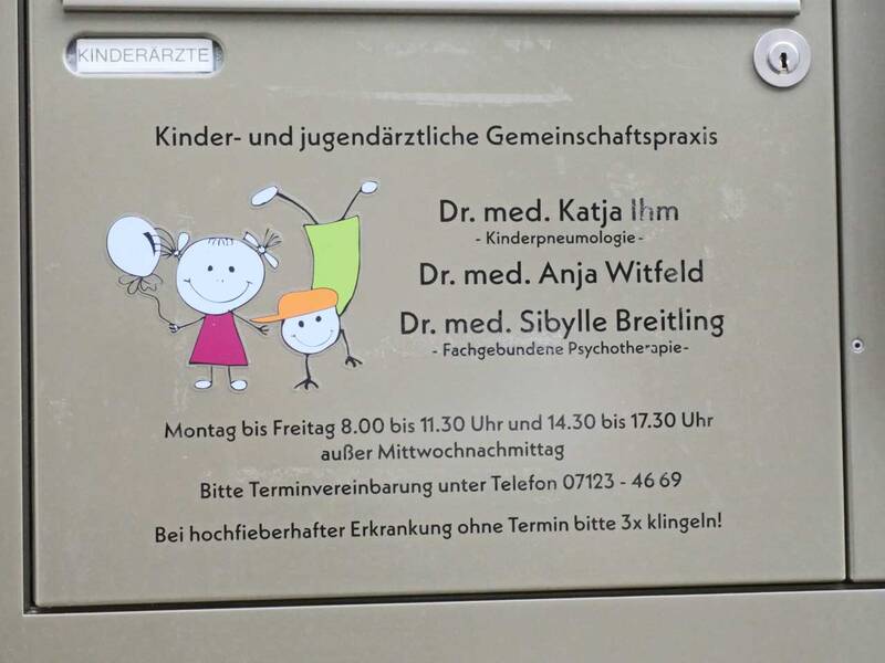 Ein Praxisschild zeigt die Öffnungszeiten und Kontaktdaten einer kinder- und jugendärztlichen Gemeinschaftspraxis mit bunten Strichmännchenillustrationen.