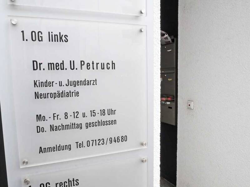 Ein Schild zeigt die Praxisinformationen eines Kinder- und Jugendarztes mit Öffnungszeiten und Telefonnummer für Termine.