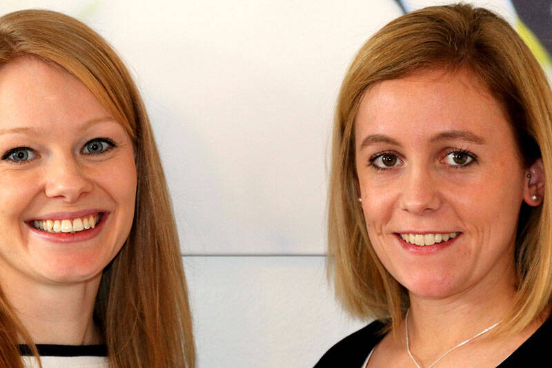 Zwei lächelnde Frauen mit blonden Haaren stehen vor einem unscharfen Hintergrund.