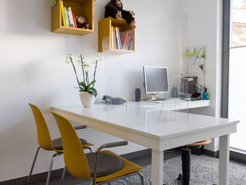 Ein heller, moderner Büroraum mit einem weißen Tisch, an dem zwei gelbe Stühle stehen, während ein Computer und Dekorationen wie eine Pflanze und Bücherregale den Hintergrund schmücken.