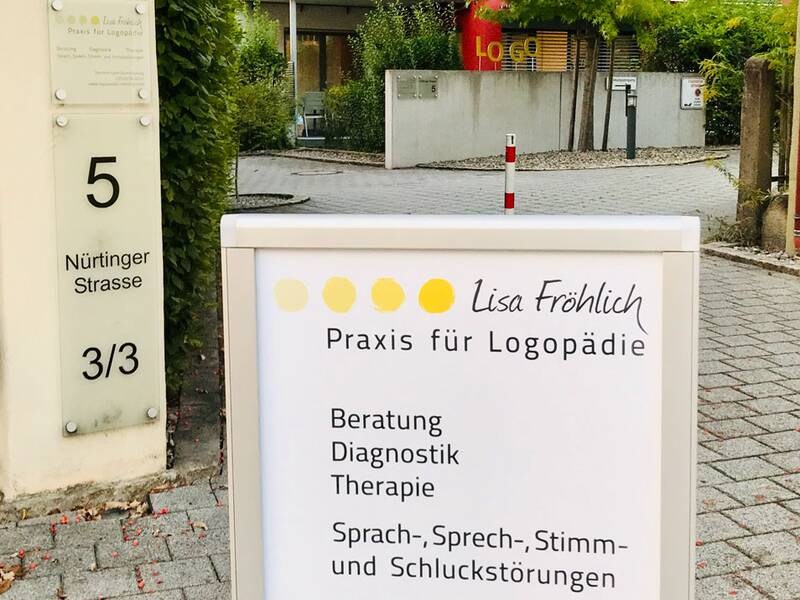 Ein Schild vor einem Gebäude informiert über eine Logopädiepraxis, die Beratung, Diagnostik und Therapie für Sprach-, Sprech-, Stimm- und Schluckstörungen bei Säuglingen, Kindern und Erwachsenen anbietet.