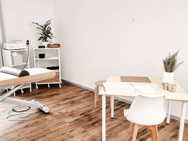 Der Raum wirkt wie ein Behandlungszimmer mit einem Holzboden, einem weißen Tisch mit Stuhl, einer Behandlungsliege und Pflanzen als Dekoration.