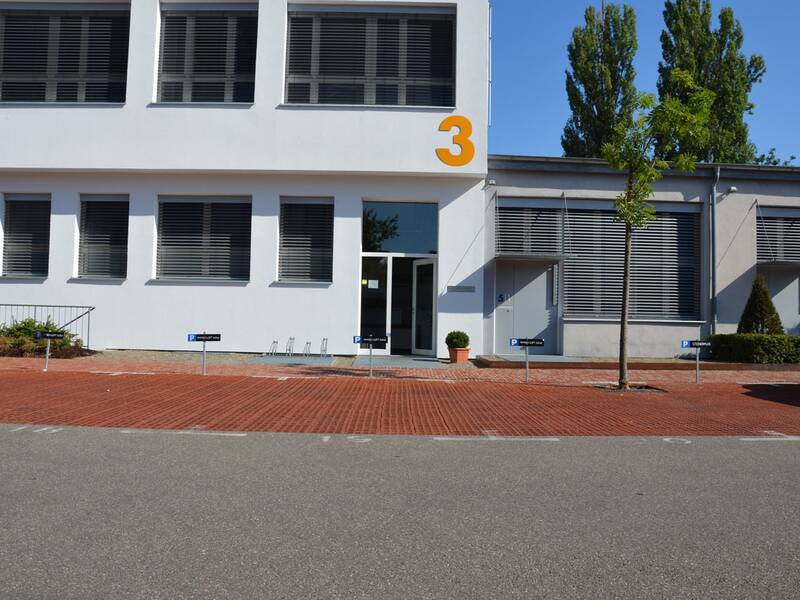 Das Bild zeigt die Frontansicht eines modernen, weißen Gebäudes mit großen Fenstern, einem zentralen Eingang, rotem Ziegelpflaster davor und der orangefarbenen Zahl „3“ an der Fassade.