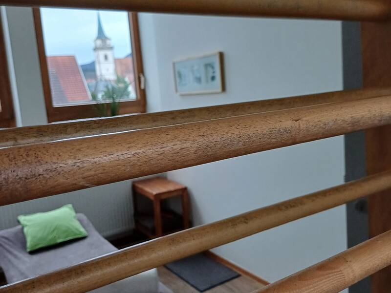 Ein Raum mit einem braunen Holzgitter im Vordergrund, dahinter ein Bett mit Kissen und ein Fenster mit Blick auf einen Kirchturm.