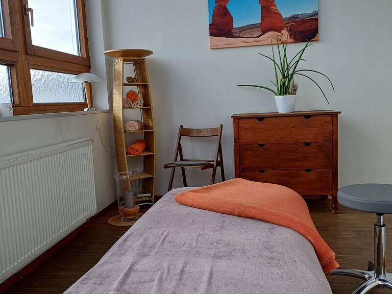 In einem hellen Raum mit einem großen Fenster steht eine Massageliege mit orangefarbenen und grauen Decken, daneben ein Regal mit Handtüchern, eine Pflanze auf einer Kommode und ein Poster an der Wand.