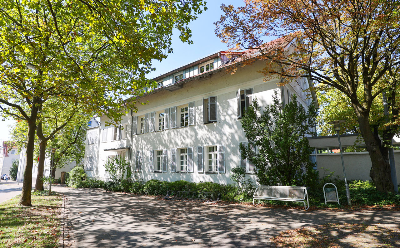 Musikschule. Ein zweistöckiges, weißes Gebäude mit Sprossenfenstern steht von Bäumen umgeben an einem sonnigen Tag.
