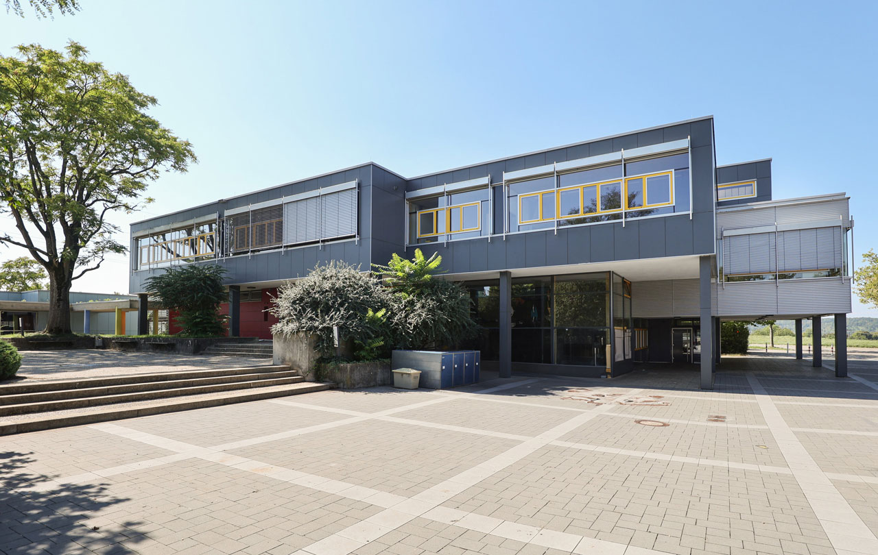 Neugreuthschule. Das Bild zeigt ein modernes zweistöckiges Gebäude mit grauer Fassade, gelben Fenstern und einer breiten Treppe, umgeben von Bäumen und Pflanzkübeln.