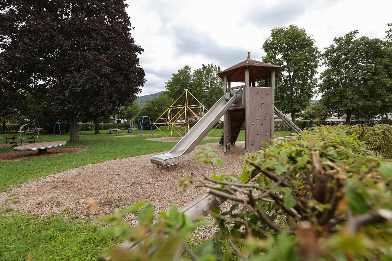 Ein großer Spielturm mit Rutsche, Kletterwand und Leiter steht auf einem Spielplatz, im Hintergrund eine gelbe Kletterpyramide und Schaukeln.