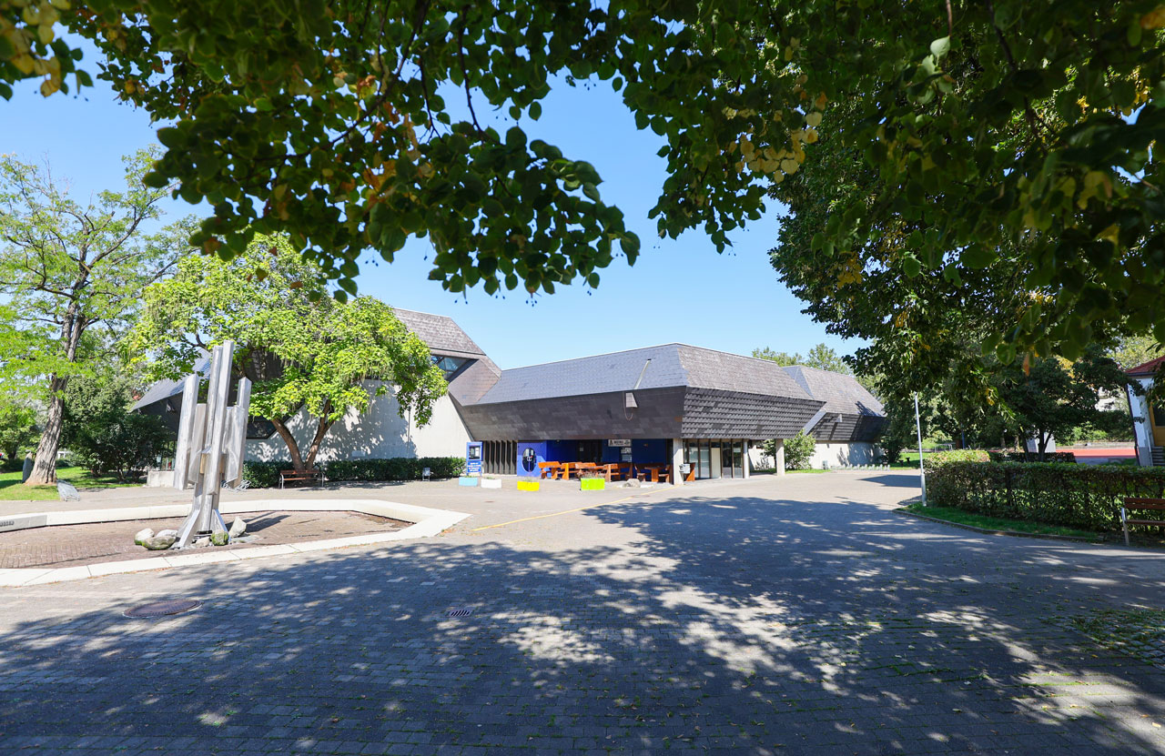 Hallenbad, Blick auf den Vorplatz mit Springbrunnen und Hallenbad im Hintergrund.